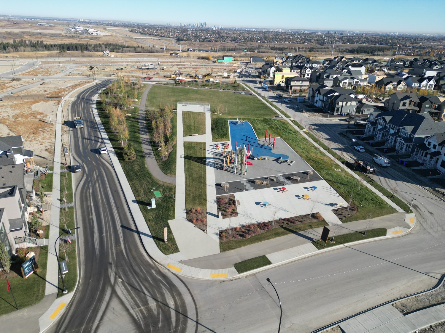 Aerial view of a suburban playground with a play structure and basketball court, surrounded by roads and residential houses. Open fields and distant city skyline are visible in the background.