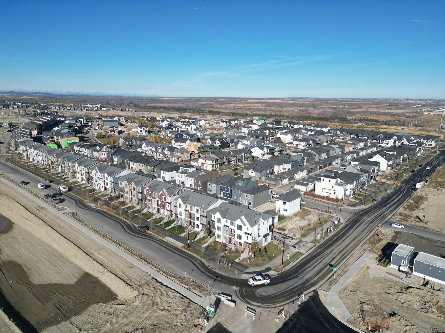 Aerial view of a suburban neighborhood with rows of houses, streets, parked cars, and surrounding open land under a clear blue sky.