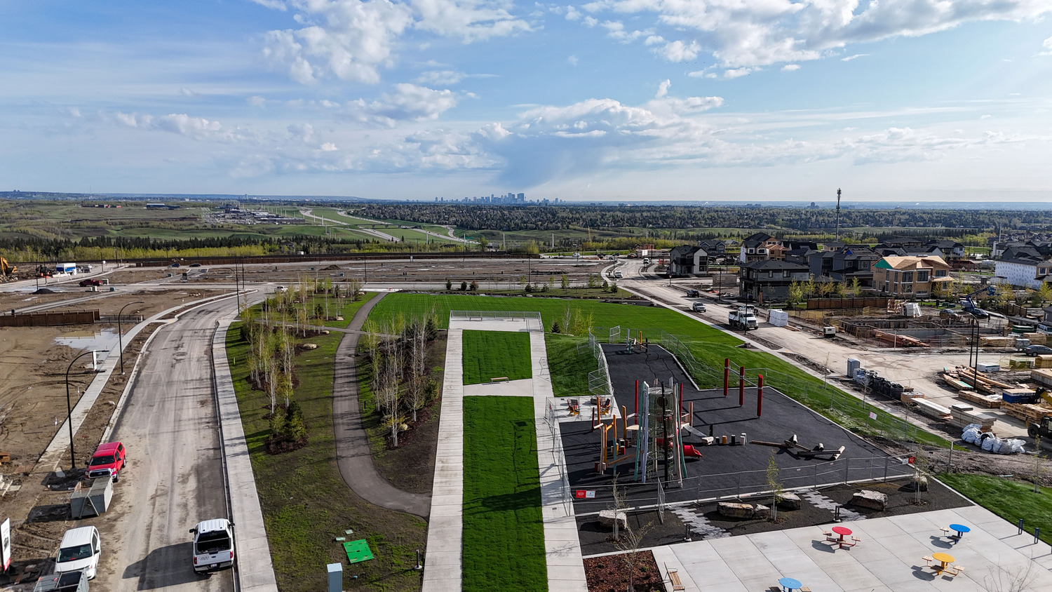 Aerial view of a suburban development area with roads, grassy fields, playground equipment, and distant city skyline. Construction vehicles and houses are visible on the right.