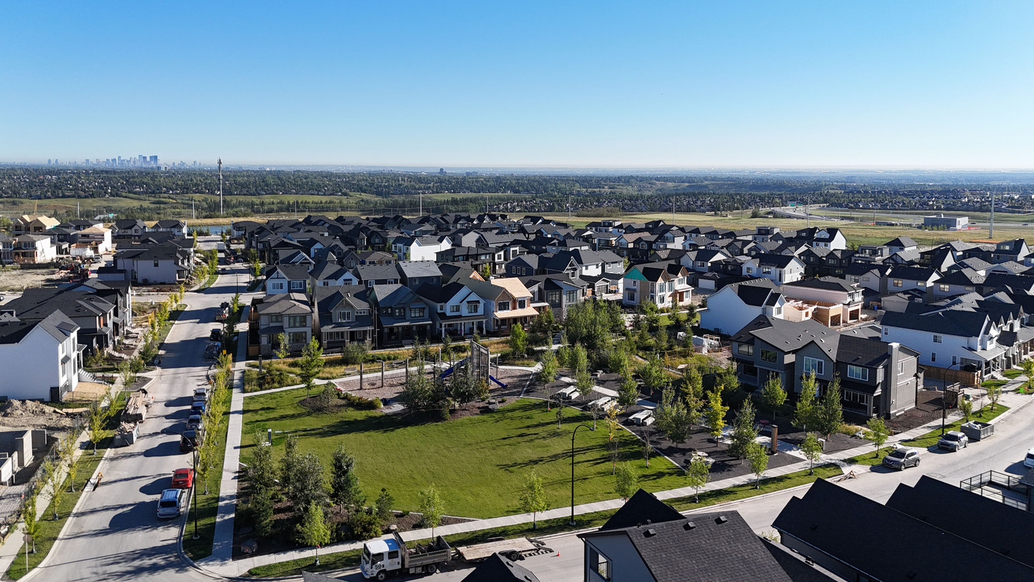 Aerial view of a suburban neighborhood with rows of houses, a central park area with trees, and a city skyline visible in the distant background under a clear blue sky.