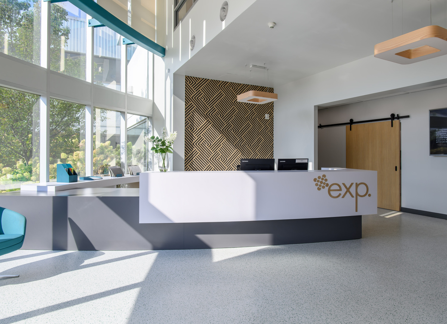 Modern office reception area with a white desk, wooden wall accents, and large windows on the left.