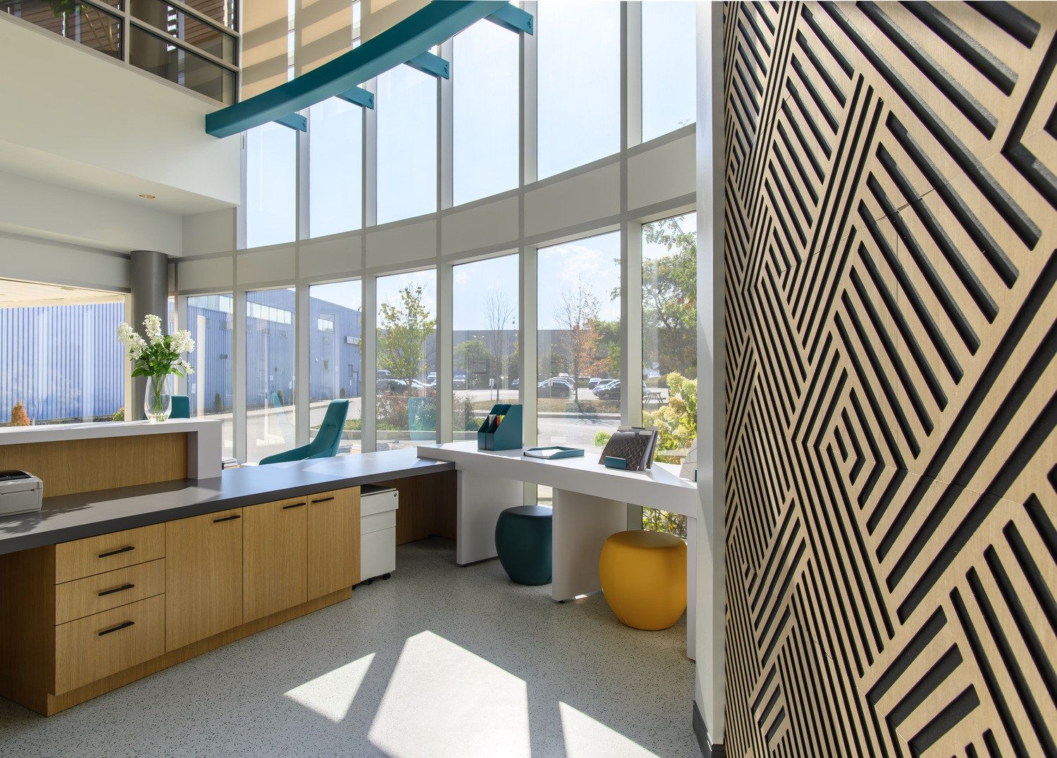 Modern office lobby featuring large windows, a decorative wooden wall, a curved desk with chairs, and potted plants.