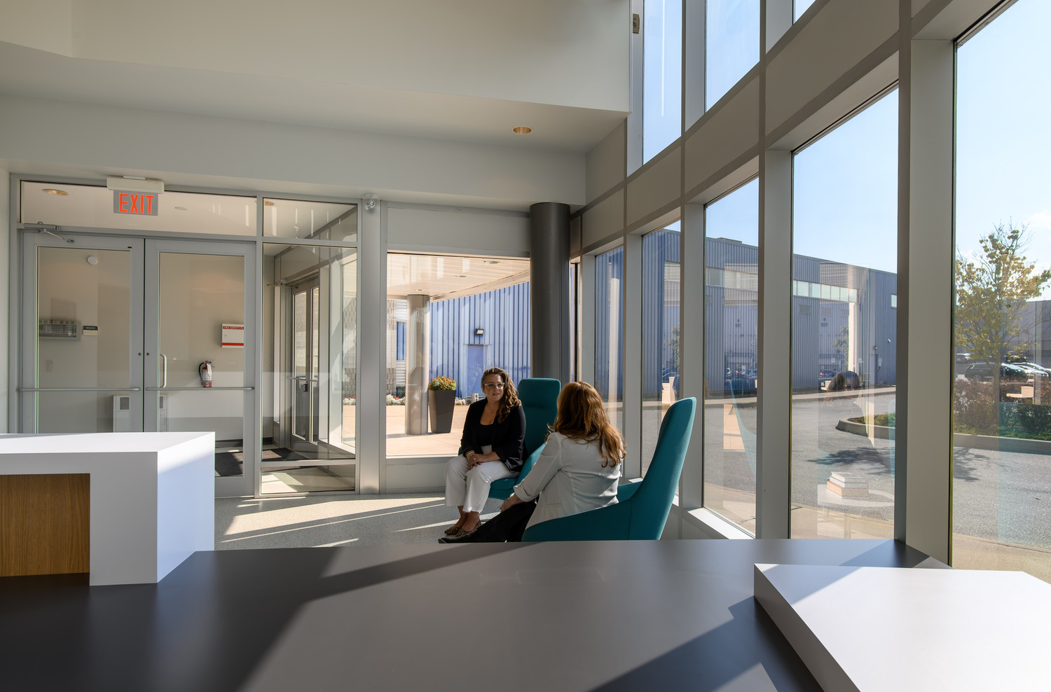 Two people seated in a modern office lobby with large windows, engaged in conversation.