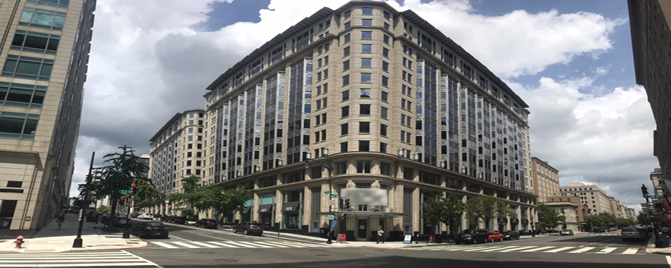 A large, multi-story office building with reflective windows stands at the intersection of two streets under a partly cloudy sky. Cars and pedestrians are visible.