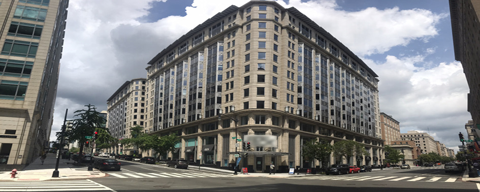 Vue grand angle d'un grand immeuble de bureaux à plusieurs étages avec des fenêtres en verre sur un coin de rue urbain, entouré de voitures et d'arbres.