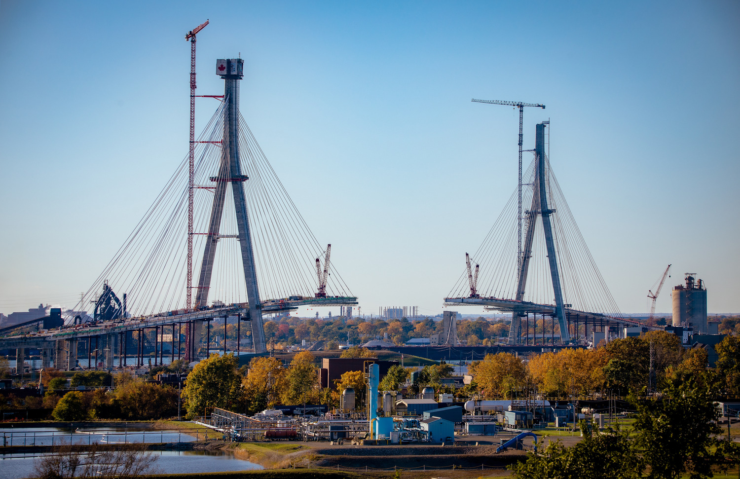 A large cable-stayed bridge under construction against a clear blue sky, with cranes and surrounding equipment visible.