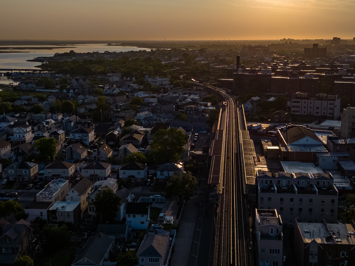 Aerial view of a railroad track running through a residential area at sunset, with houses on either side and a body of water in the background.