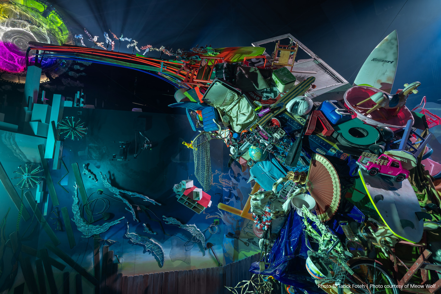 A vibrant indoor art installation featuring a complex structure made of colorful objects, with various shapes and textures, under dynamic lighting.