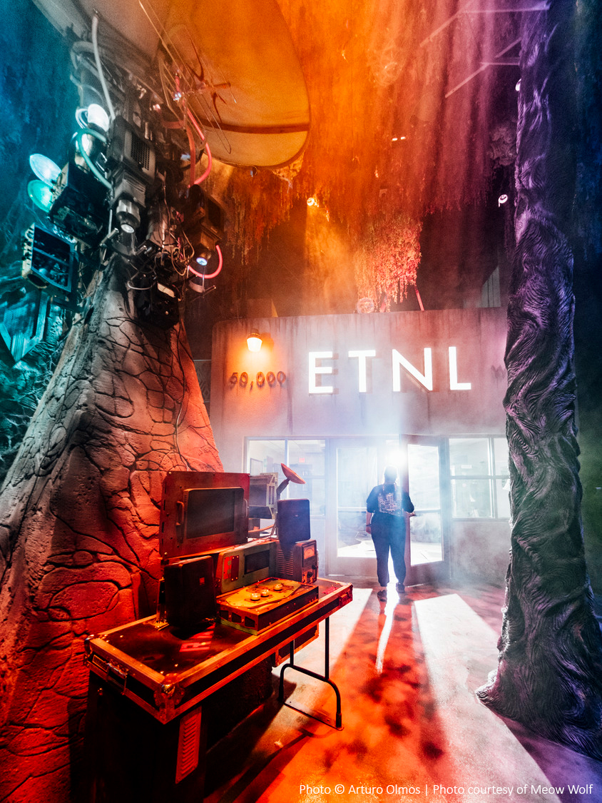 Colorful, misty interior with electronic equipment and an illuminated ETNL sign. Trees and lights frame the scene, with a person standing near the entrance.