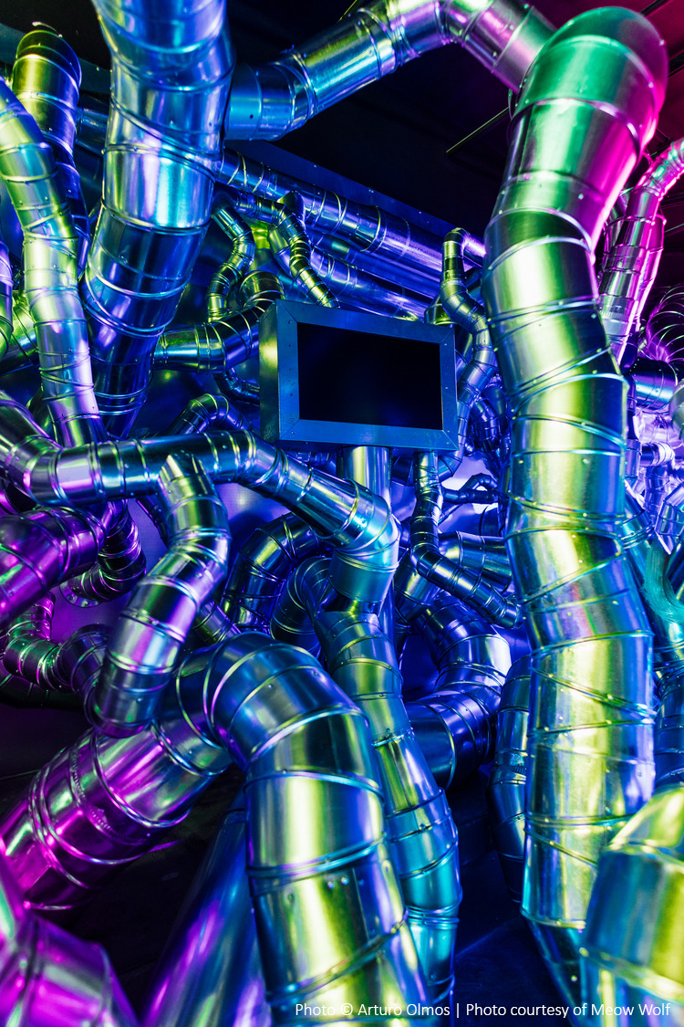 A tangle of metallic pipes in vibrant lighting with a small rectangular screen in the center.