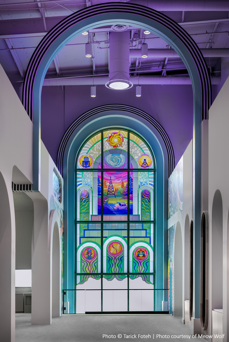 Colorful stained glass window in an arched frame, depicting abstract designs, is illuminated in an art gallery with purple lighting.