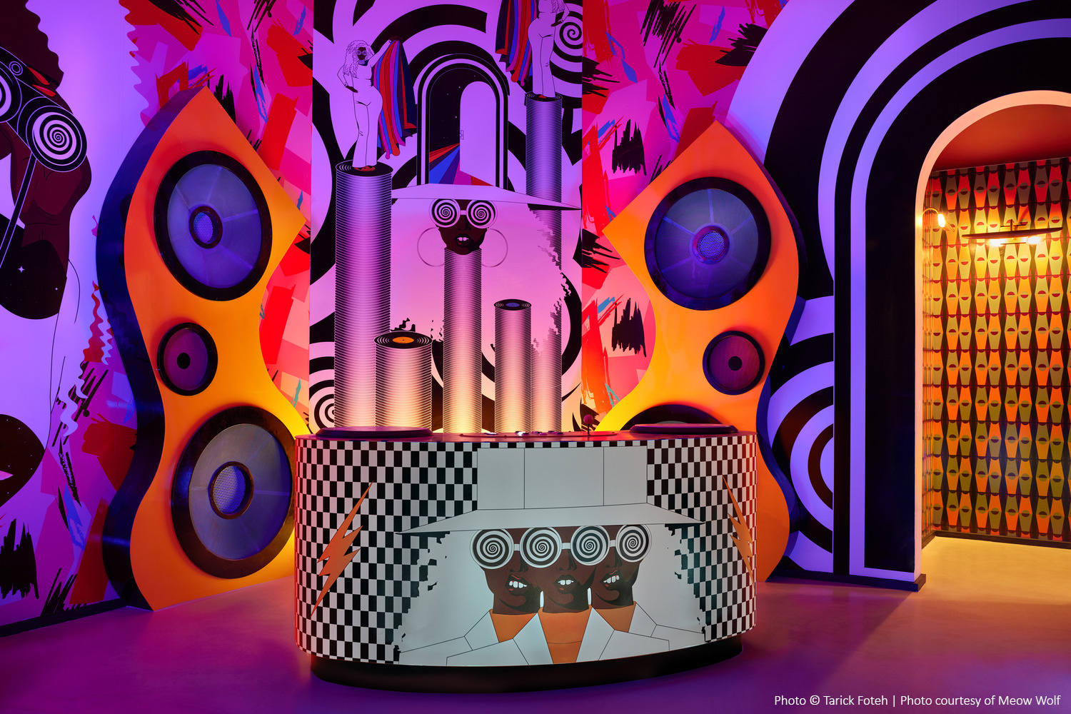 Colorful art installation featuring large speakers, abstract patterns, and neon lighting, with a stylized DJ booth in the center.