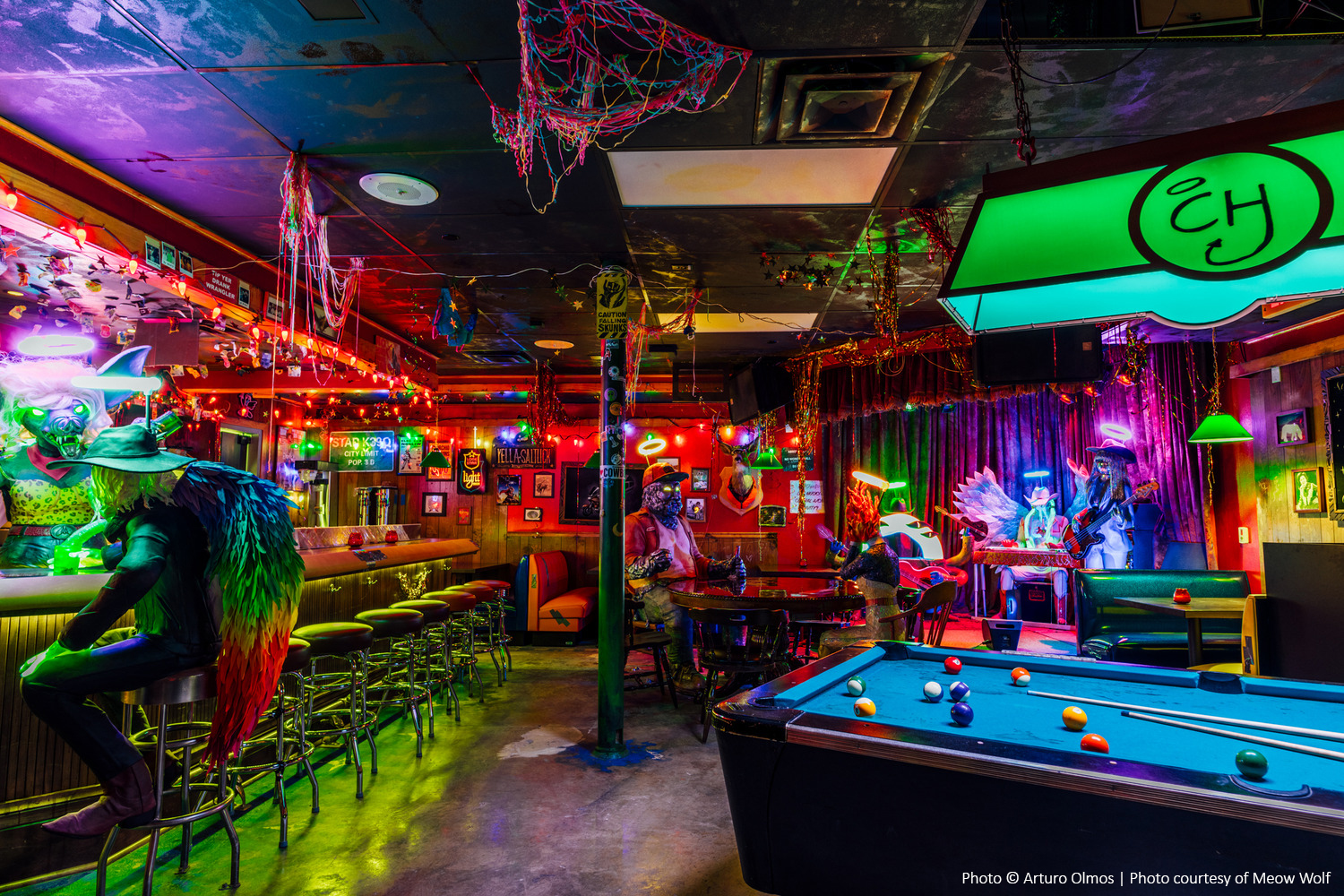 Vibrant bar with neon lights, colorful decor, masked figures, pool table, and lively atmosphere.