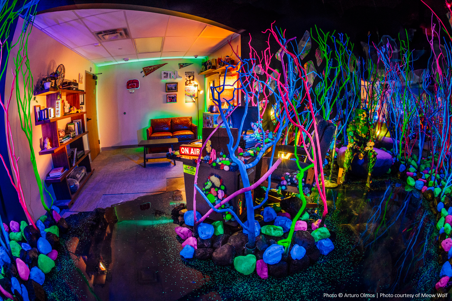 A room with neon-lit decor features colorful glowing branches, a couch, and a wall-mounted TV. The scene is vibrant and surreal, blending living room elements with artistic installations.