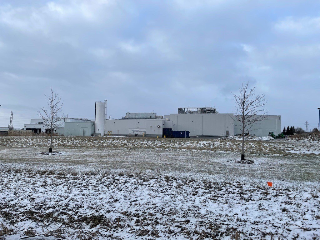 A large industrial building stands on a snowy landscape with a cloudy sky. Sparse trees are visible in the foreground.