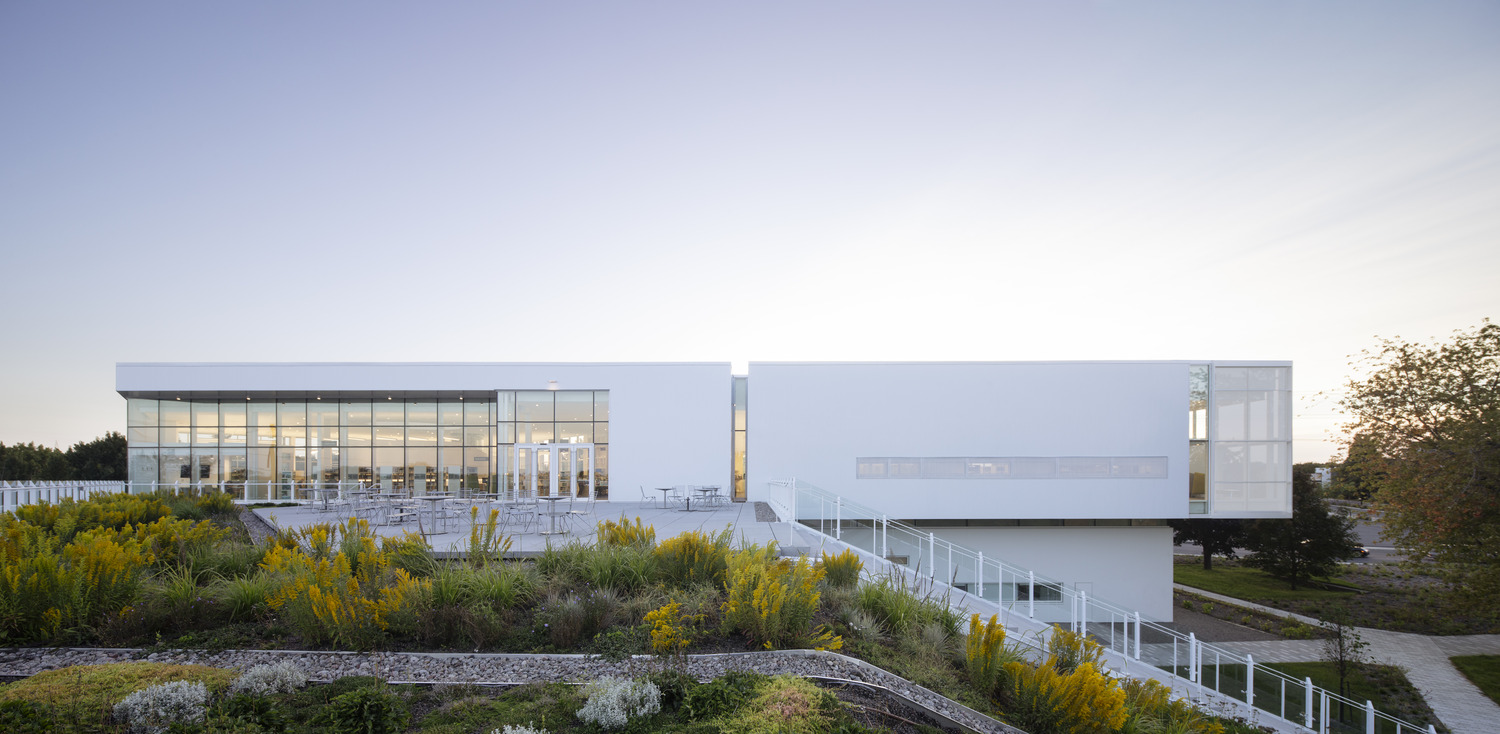 Modern white building with large windows, surrounded by landscaped greenery and outdoor seating under a clear sky.