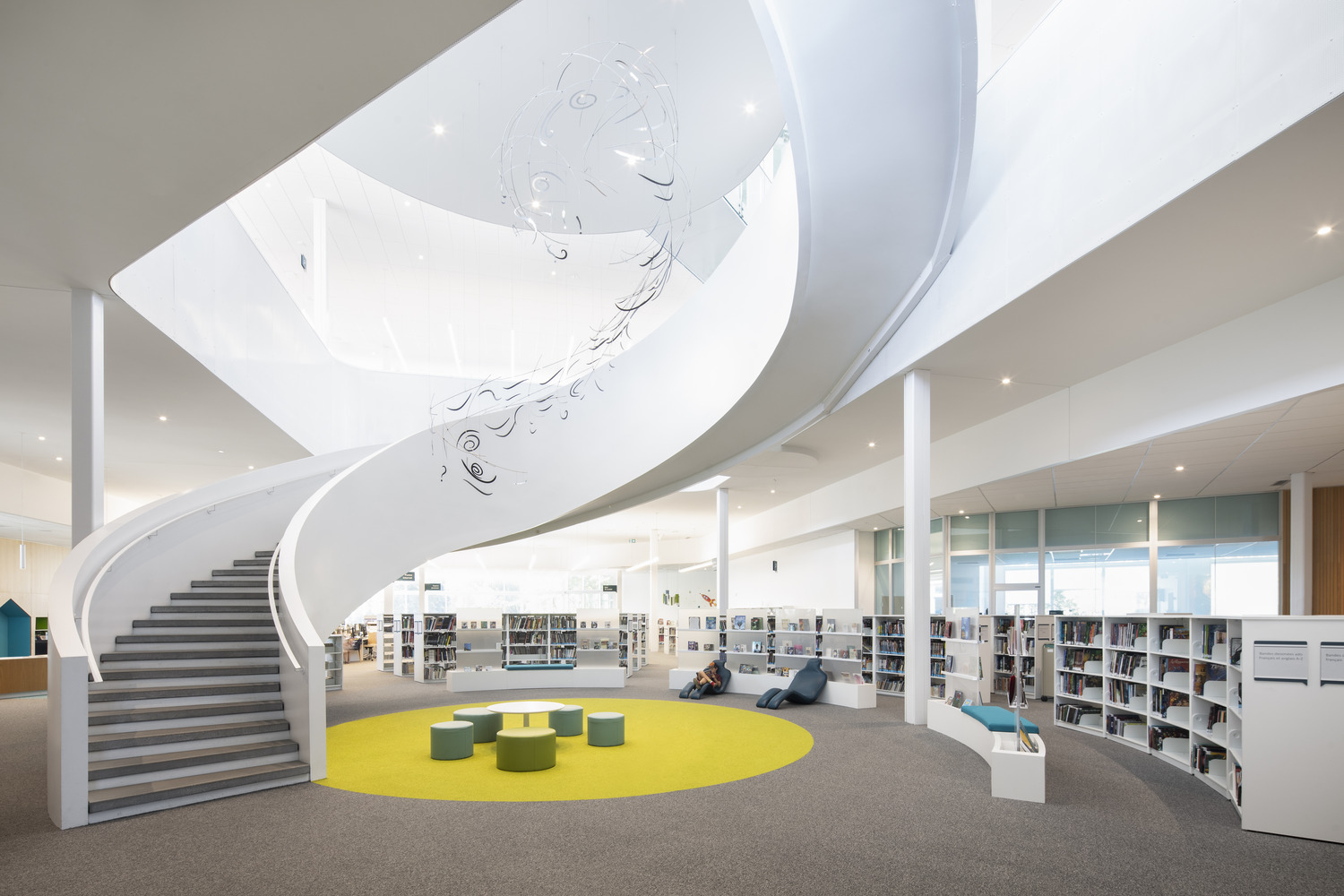 A modern library interior with a spiral staircase, circular yellow rug, seating area, bookshelves, and white walls.