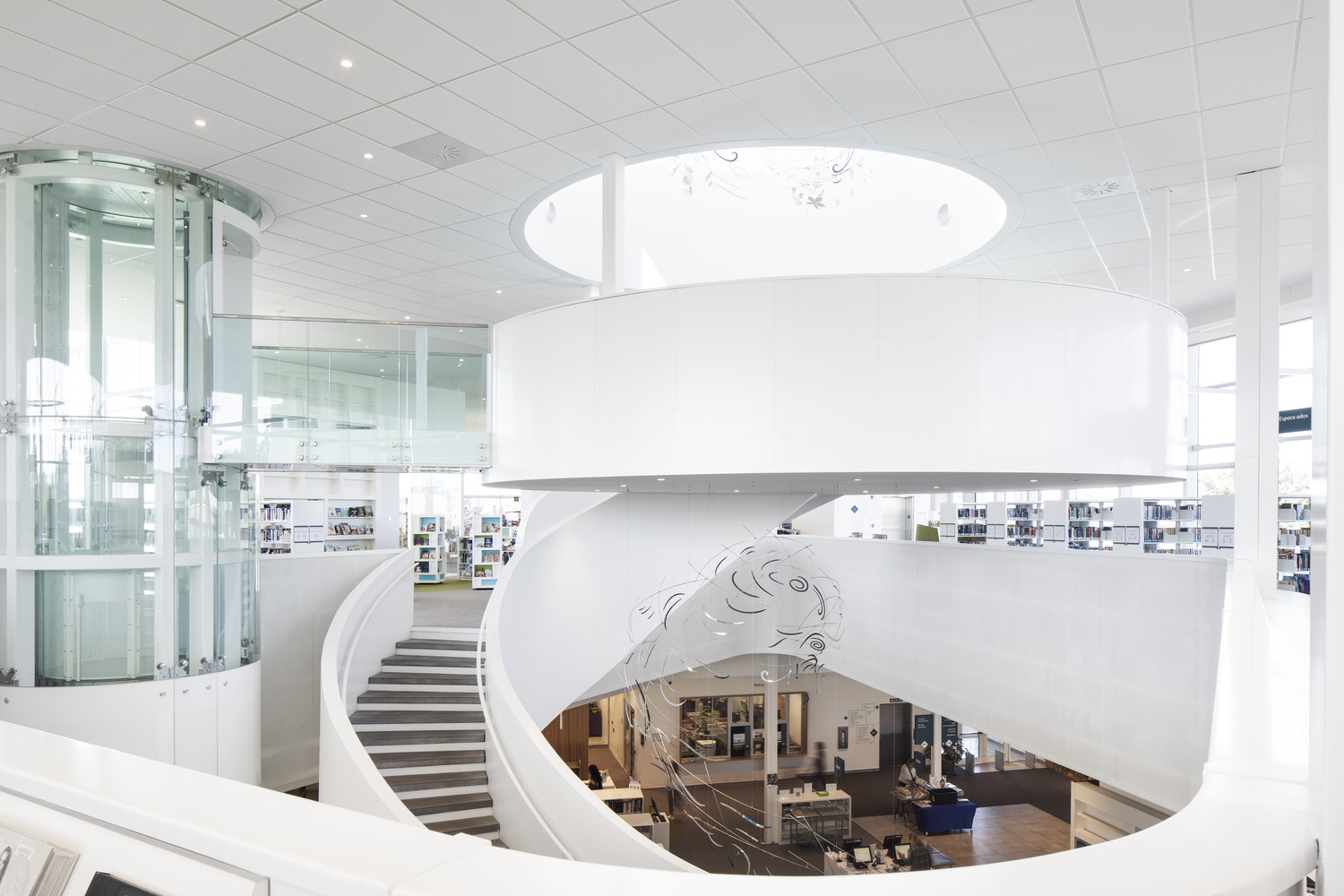 Modern library interior with a large spiral staircase, glass elevator, and shelves of books. There is ample natural light and white architectural elements.