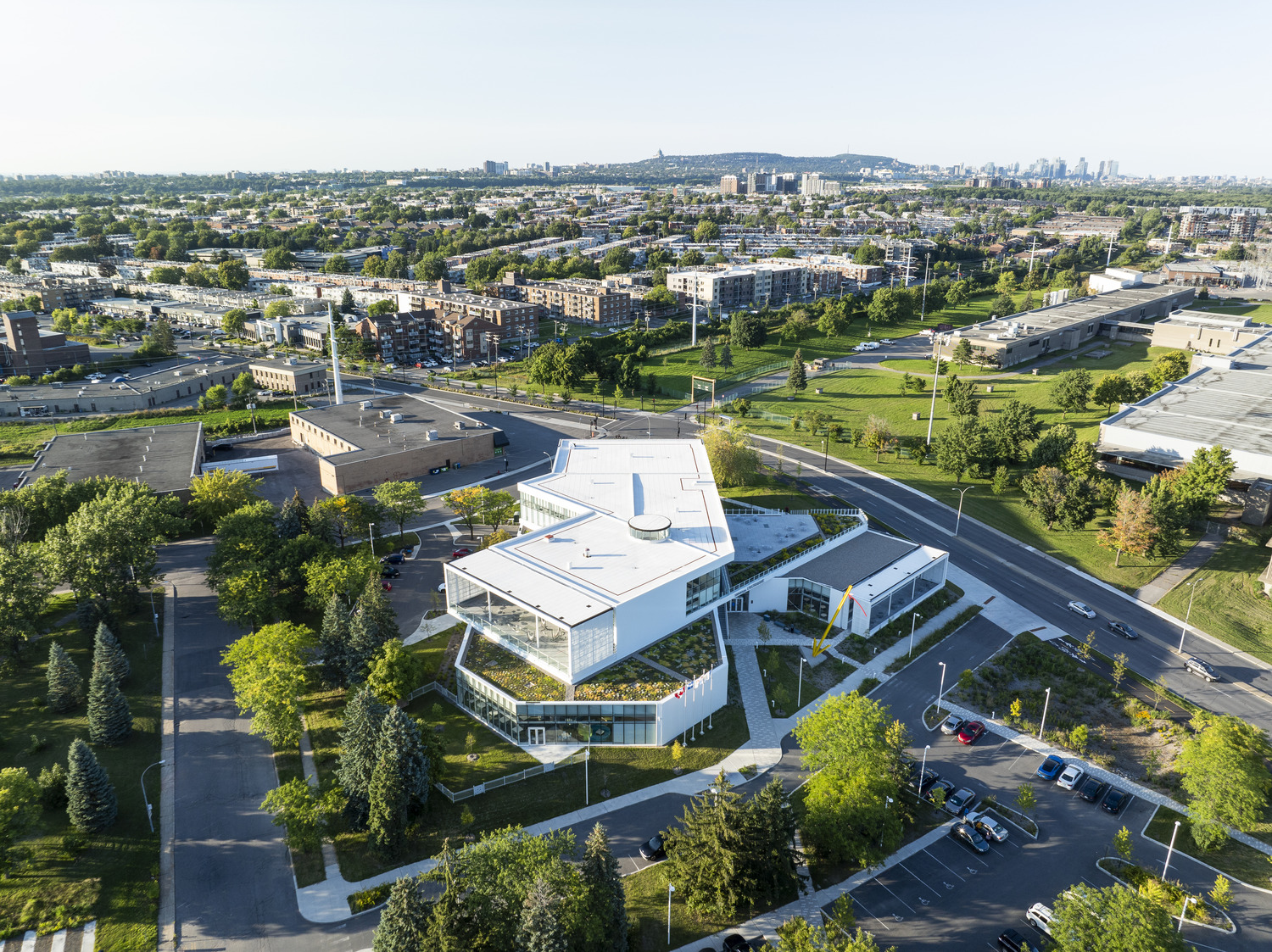 Aerial view of a modern, multi-story building with a white roof, surrounded by greenery and roads, in an urban area with a cityscape in the distance.