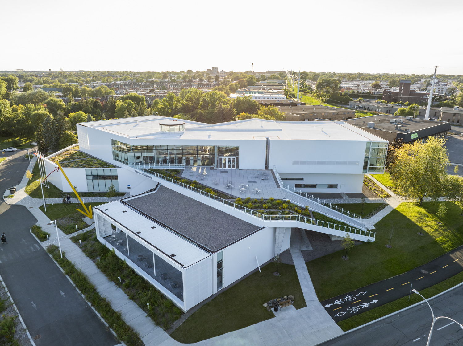 Modern white building with angular design, surrounded by greenery and roads. Rooftop terrace visible.