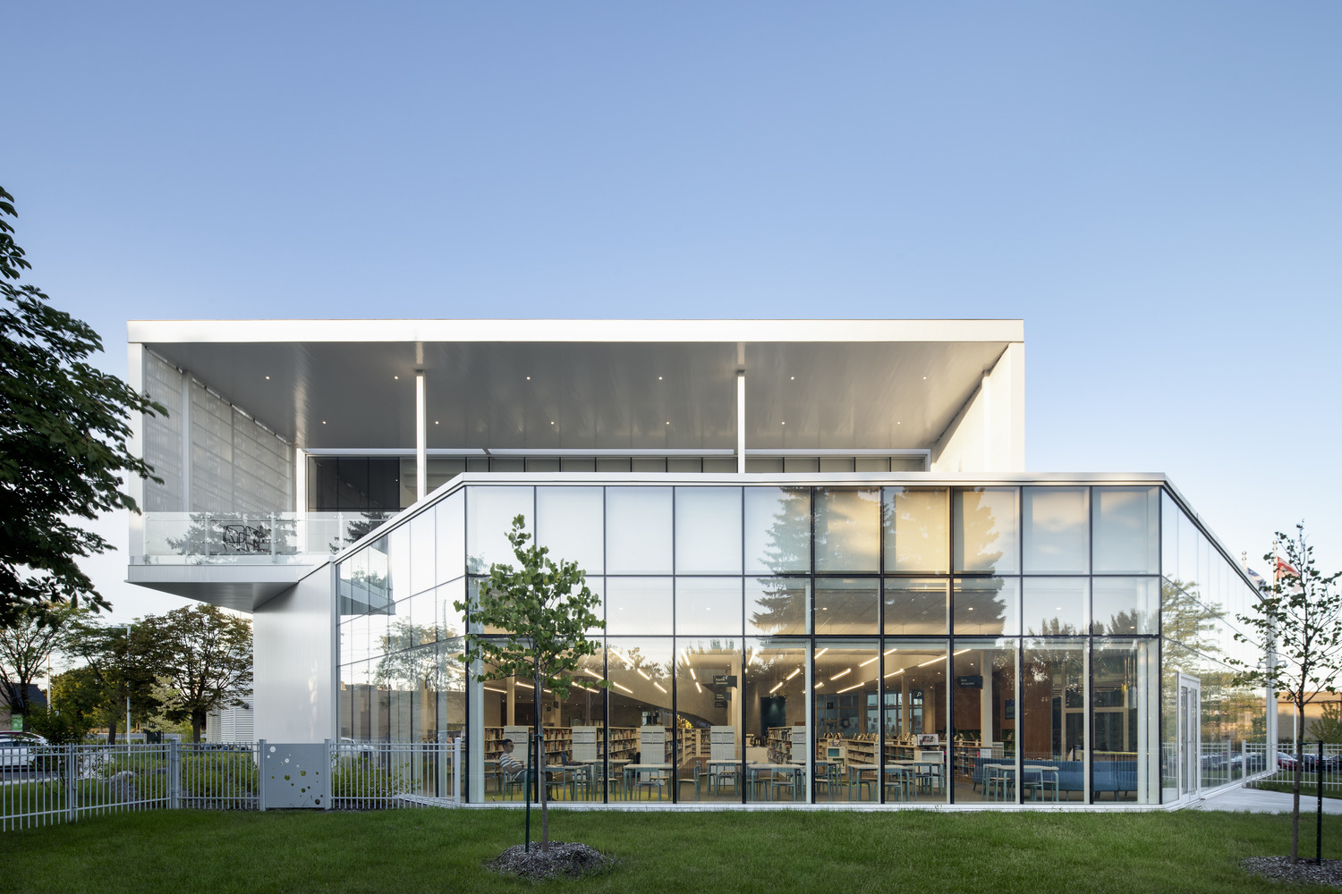 Modern two-story glass building with open upper deck, surrounded by grass and trees under a clear blue sky.