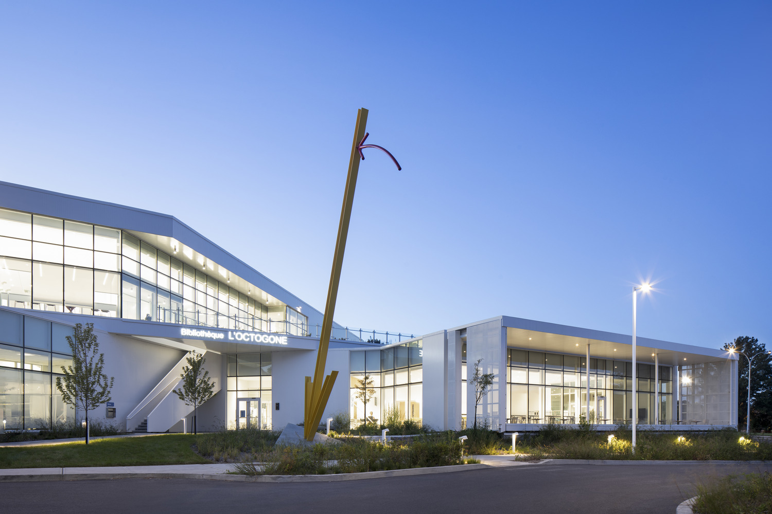 Modern library building with large glass windows and an abstract sculpture in front, under a clear blue sky.