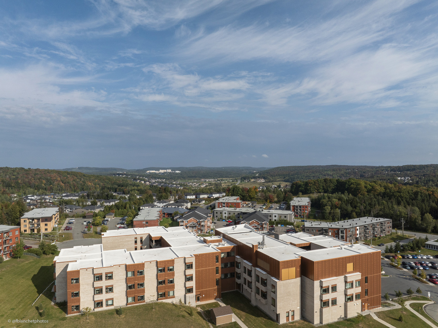 Aerial view of a multi-story brick building surrounded by greenery and other structures, with a vast landscape and scattered clouds in the background.