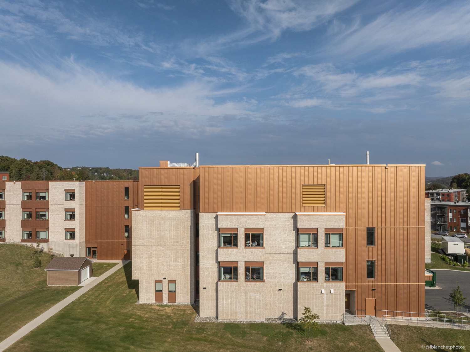 A modern building with copper-colored exterior panels and large windows, surrounded by grass and a paved pathway under a partly cloudy sky.