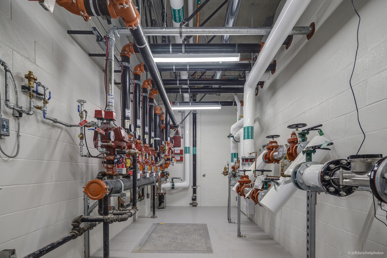 Industrial room with pipes and valves along white walls, including red and green components. Overhead lighting illuminates the space.