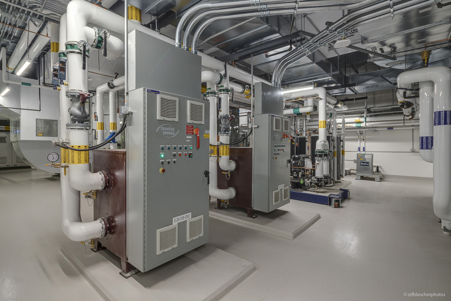 Industrial room with large gray machinery, pipes, and control panels. Ceiling shows exposed ducts and cables. Floor is smooth and light-colored.