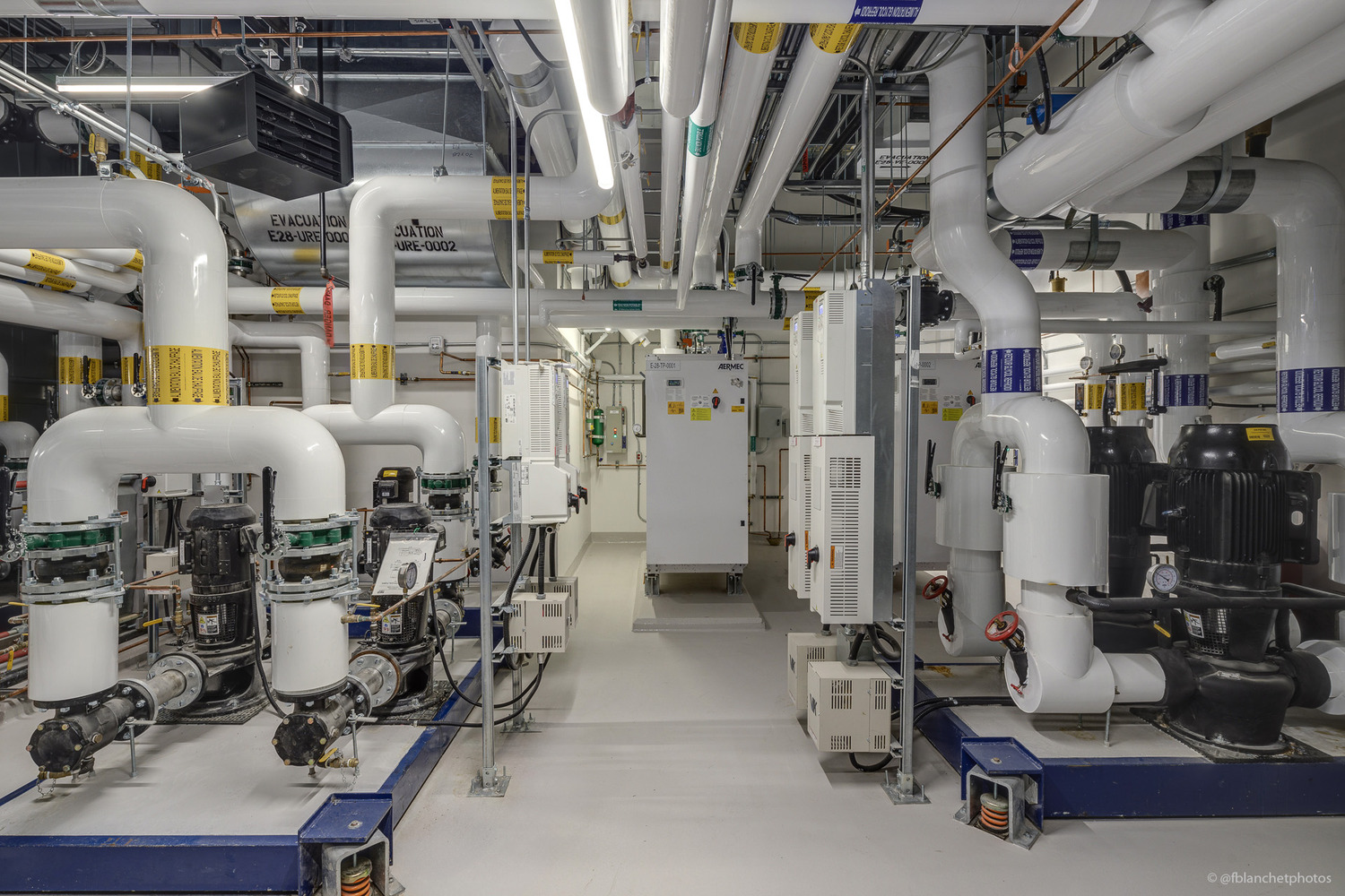 Industrial room with large white pipes, valves, machinery, and control panels. Bright overhead lighting and a clean, organized layout feature prominently.