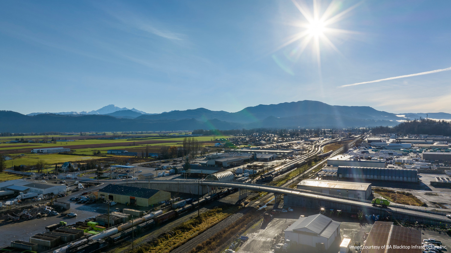 Aerial view of a town with a railway yard, buildings, and fields under a clear blue sky. The sun shines brightly over distant mountains.