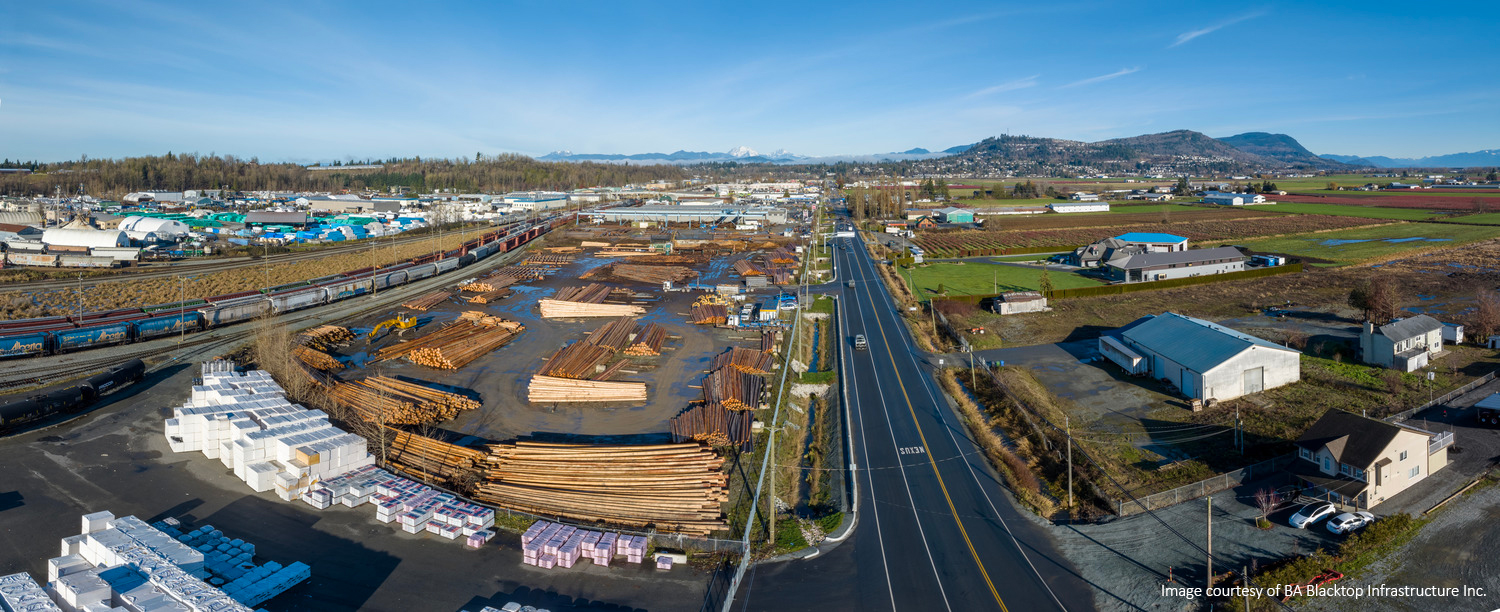 Aerial view of a lumber yard with stacks of logs and materials, adjacent to a road. Farmland and distant hills are visible under a clear blue sky.