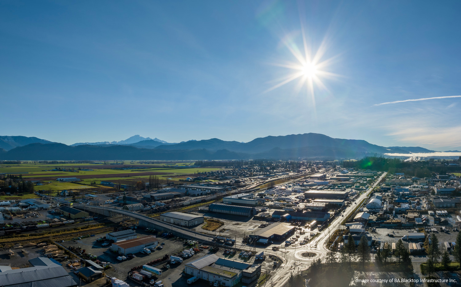 Aerial view of a town with industrial buildings, roads, and surrounding fields under a bright sun in a clear blue sky, with mountains in the background.