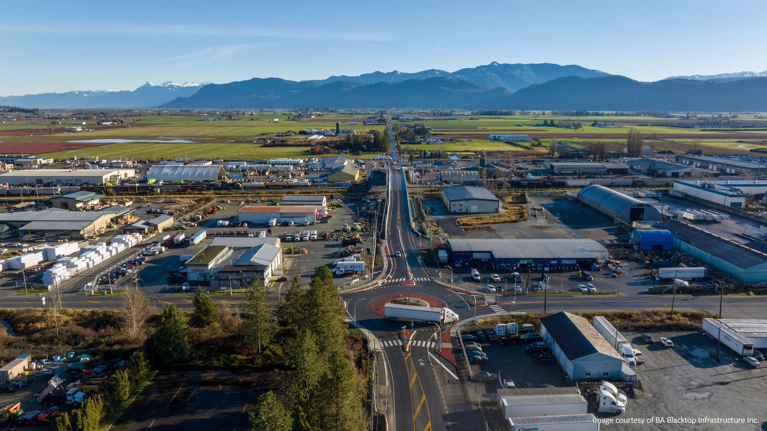 Aerial view of an industrial area with warehouses and farmland, set against a backdrop of distant mountains under a clear sky. A road runs through the center of the image.