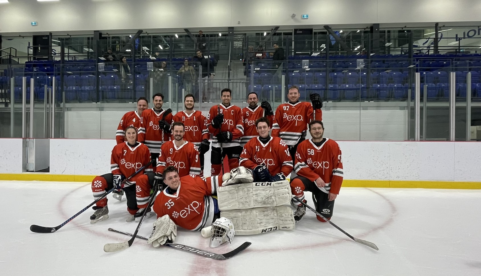 Une équipe de hockey en maillot rouge pose sur la patinoire. Onze joueurs, certains à genoux, et deux gardiens de but se tiennent devant. Les gradins à l'arrière-plan sont pour la plupart vides.