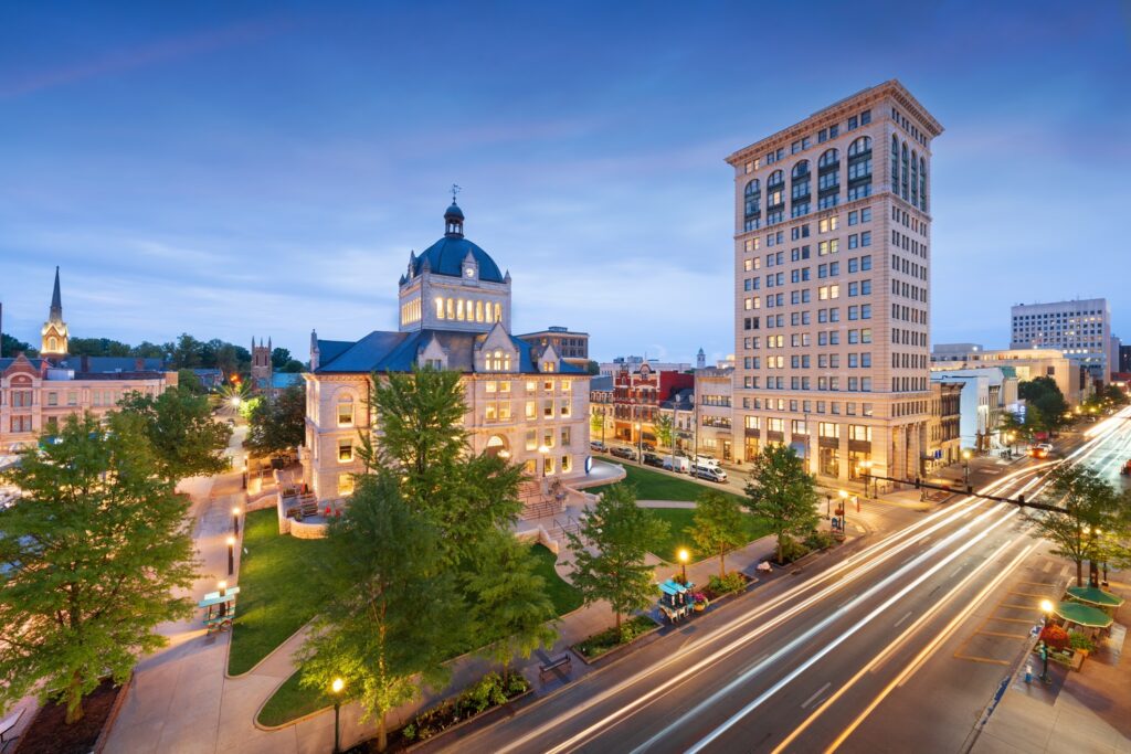 Cityscape at dusk showcasing a historic courthouse and tall building, with illuminated streets bustling with transportation. Surrounded by trees, this vibrant scene captures the essence of a modern metropolis preparing for NASTO 2025.