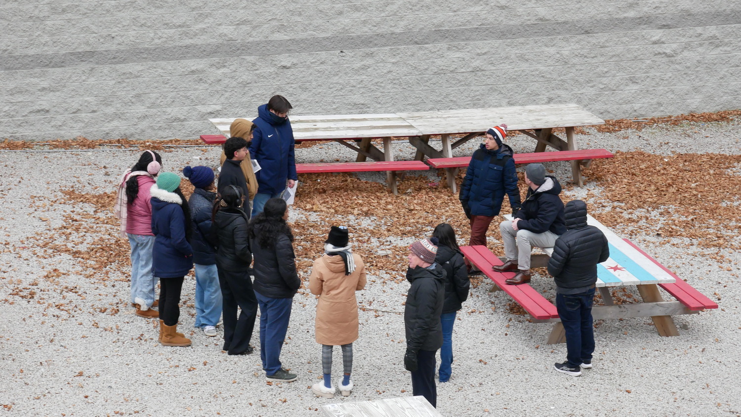 A group of people in winter clothing are standing and sitting on picnic tables in an outdoor area covered with fallen leaves.