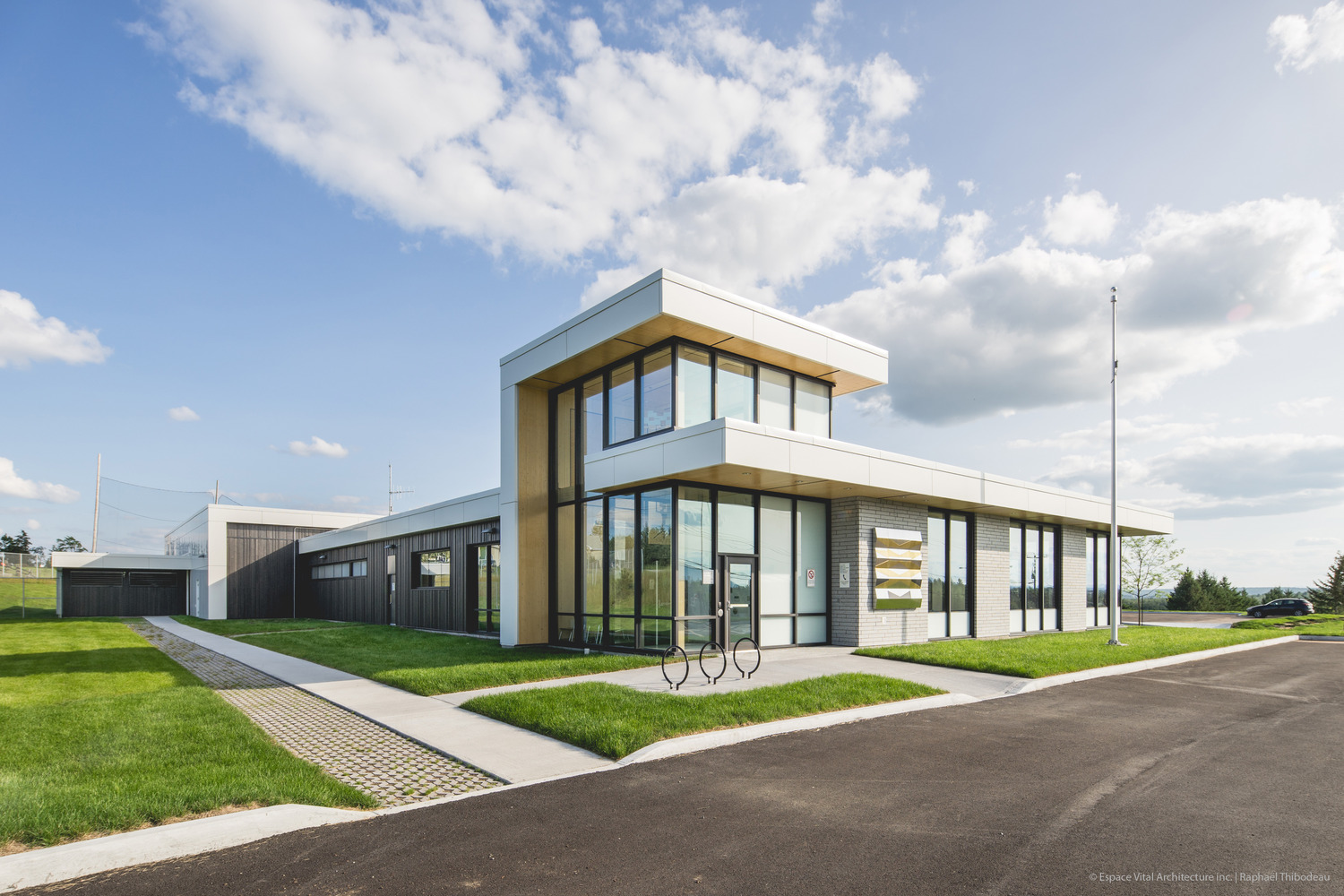 Modern single-story police station building with large windows and a flat roof, surrounded by grass and a paved driveway, under a partly cloudy sky.