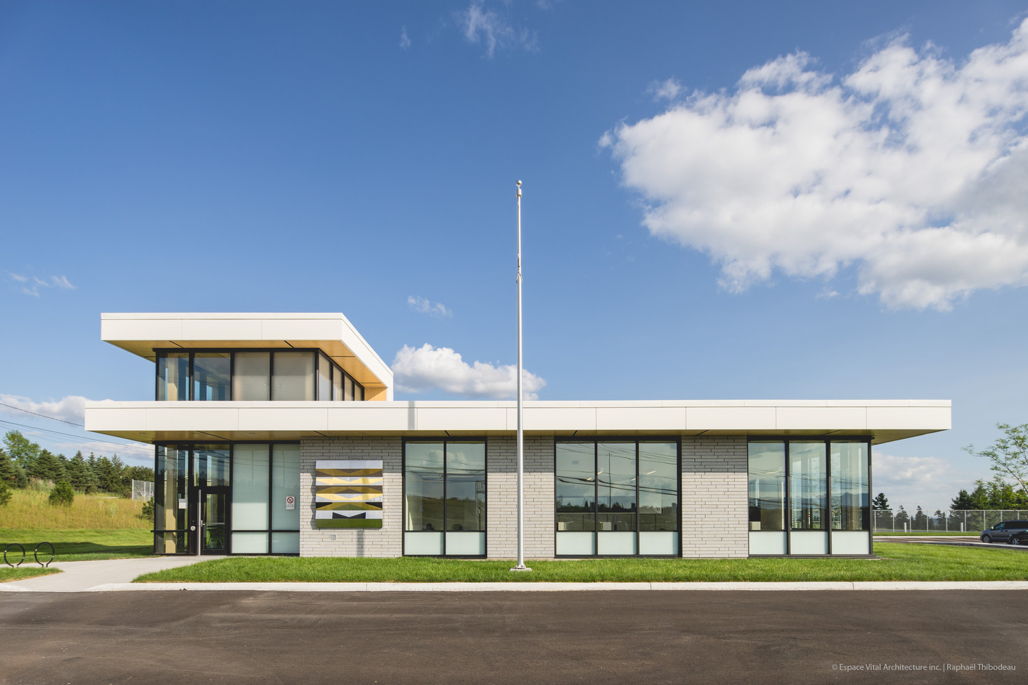 Modern, single-story police station building with large windows, adjacent tower section, surrounded by grass, under a blue sky with clouds.