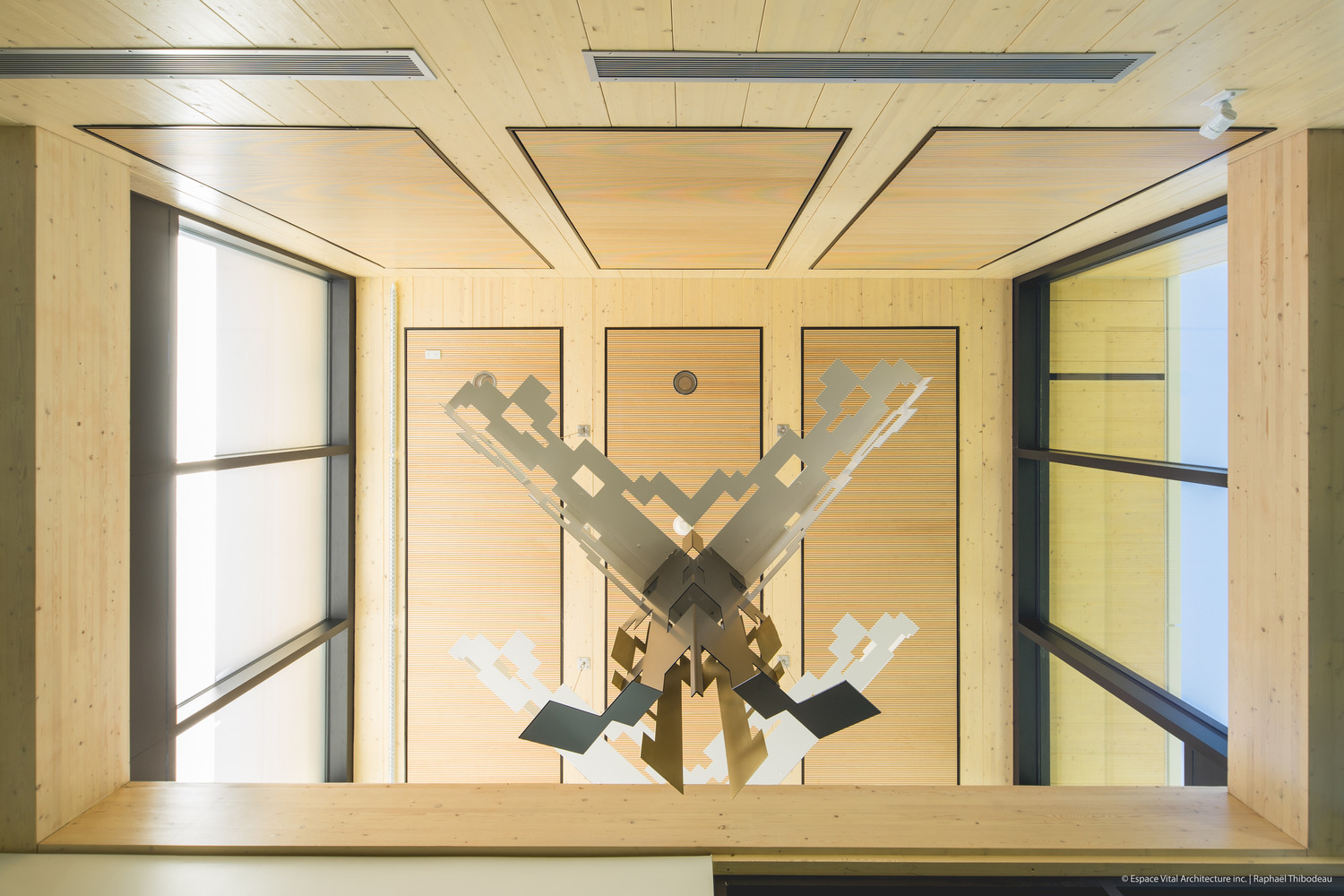 Ceiling view inside new police station featuring a modern, abstract sculpture with geometric metal elements and wood paneling, flanked by large windows.