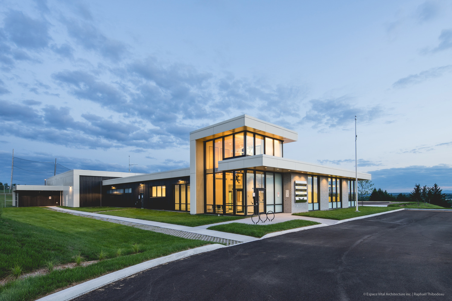 Modern police station building with large glass windows and a flat roof, surrounded by grass and a paved road, set against a twilight sky.