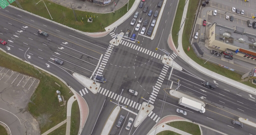 An aerial view of a bustling Canadian intersection showcases multiple lanes, crosswalks, and vehicles. Sidewalks and green areas envelop the roads, capturing the vibrant scene reminiscent of the dynamic discussions at the ITE Canada 2025 Annual Conference.