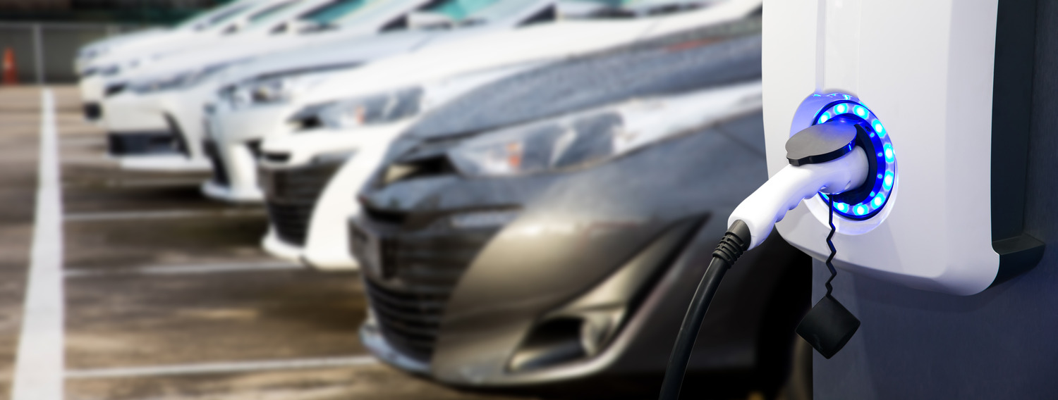 Electric vehicle charging at a station with a row of parked cars in the background.