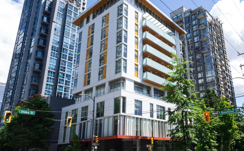 Nestled at a street corner, the modern multi-story Covenant House boasts a robust building envelope. It stands amidst towering skyscrapers, with traffic lights and signs for Drake St and Seymour St defining the vibrant foreground.