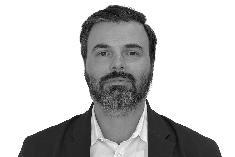 Bearded man with short hair, wearing a suit jacket over a shirt, poses against a plain white background.